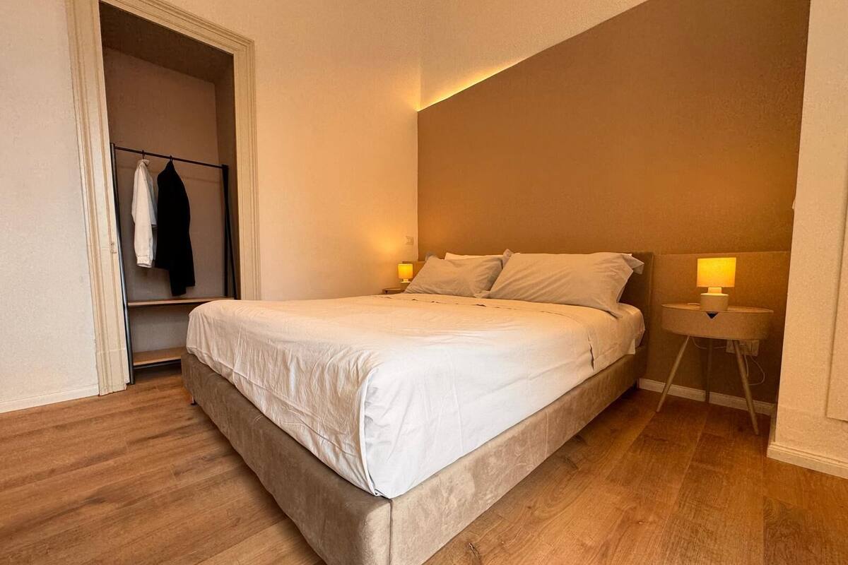 A bedroom with a bed, bedside lamps, a wooden floor, and a closet with hanging clothes.