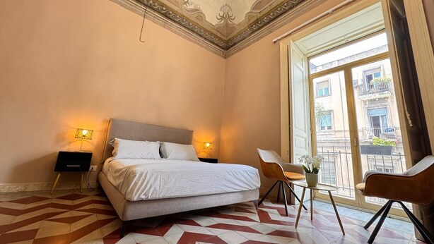 A hotel room with a bed, two bedside lamps, a chair, a small table, and a view of the outside balcony. - Dimore del Plebiscito B&B (Catania)