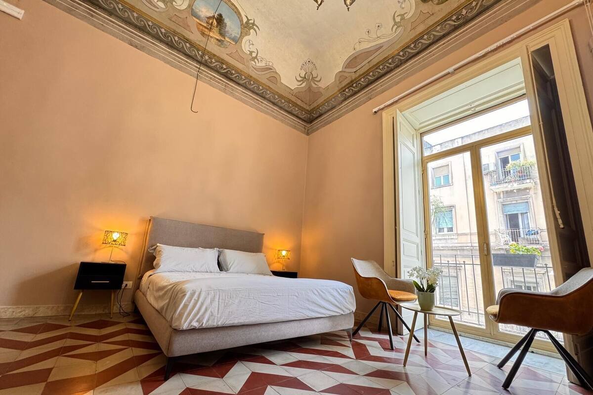 A hotel room with a bed, two bedside lamps, a chair, a small table, and a view of the outside balcony.