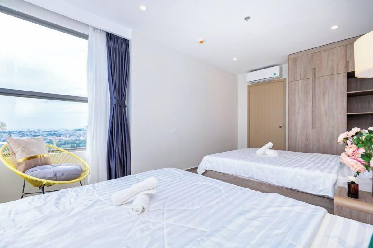 A hotel room with two beds, a large window with curtains, a yellow chair, and a view of the city.