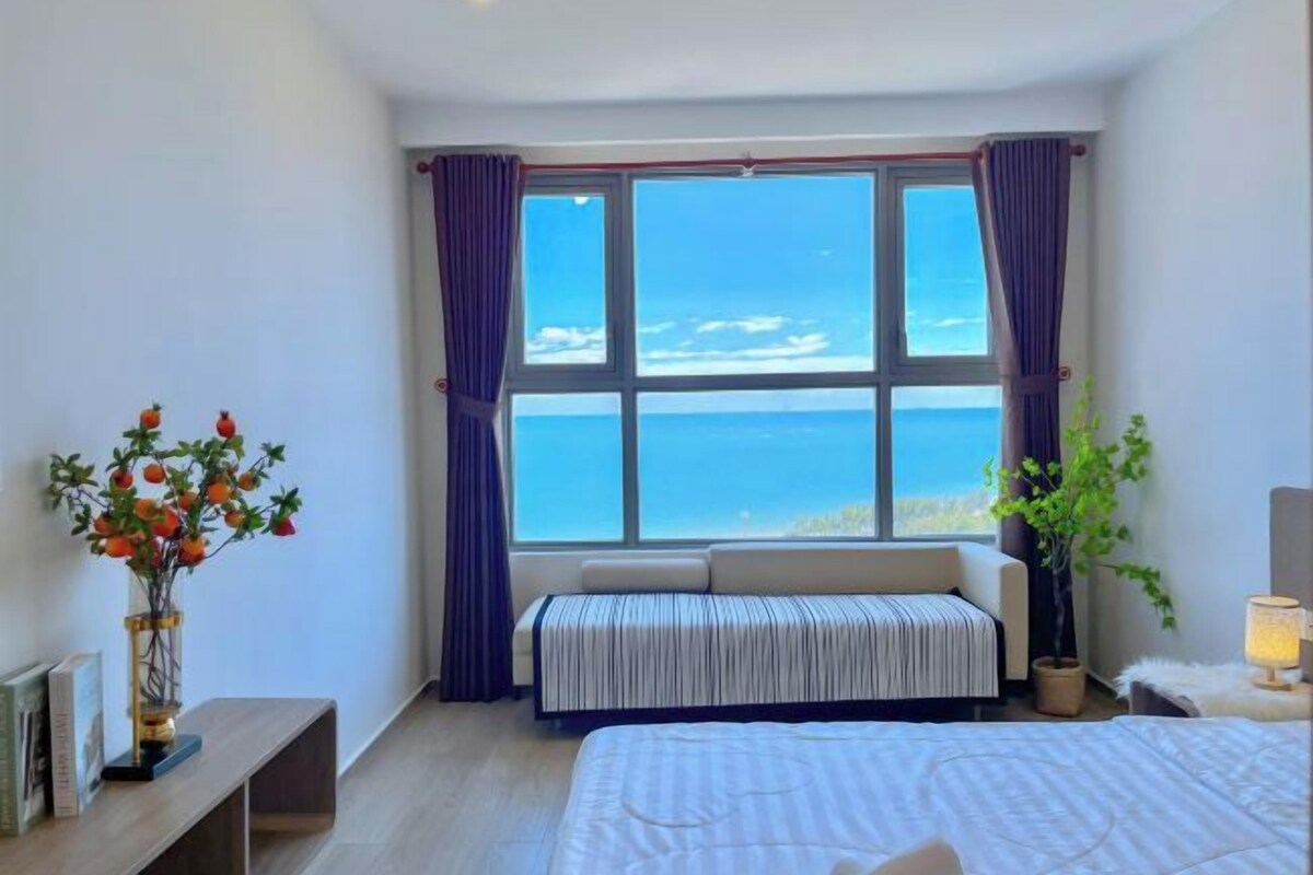 A bedroom with a large window, a bedside table with a vase of flowers, a bench, and a bedside lamp.