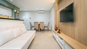 Una sala de estar moderna con un sofá, una mesa de comedor con sillas, un televisor montado en la pared y una zona de cocina con armarios y electrodomésticos.