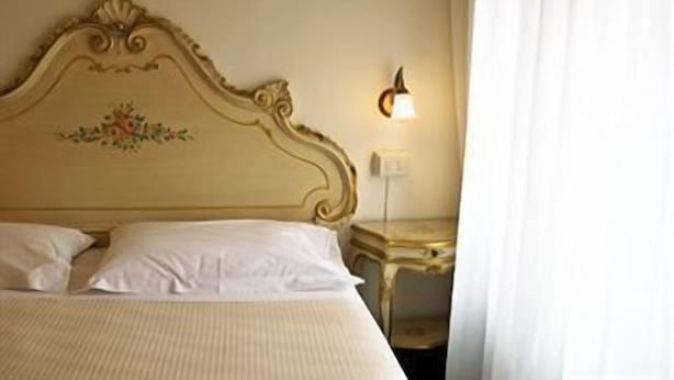 A bed with white linens, a decorative headboard, a bedside table, and a wall-mounted light. - The Venice Times (Venice)