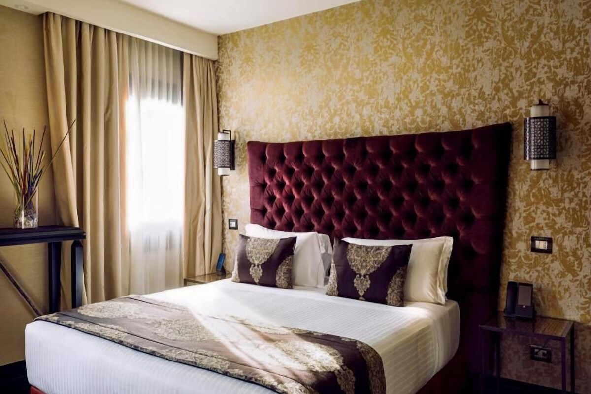 A hotel room with a large bed, a headboard, two wall sconces, a bedside table, and a window with curtains.
