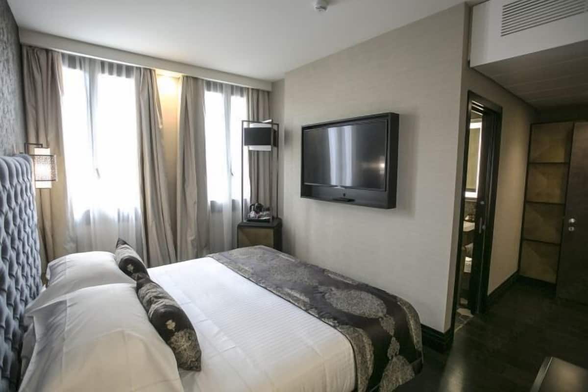A hotel room with a bed, a flat-screen TV mounted on the wall, a bedside table, and a door leading to another room.