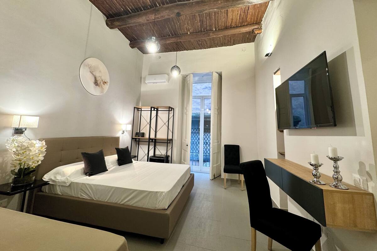 Een slaapkamer met een bed, nachtkastje, lamp en een aan de muur bevestigde televisie.