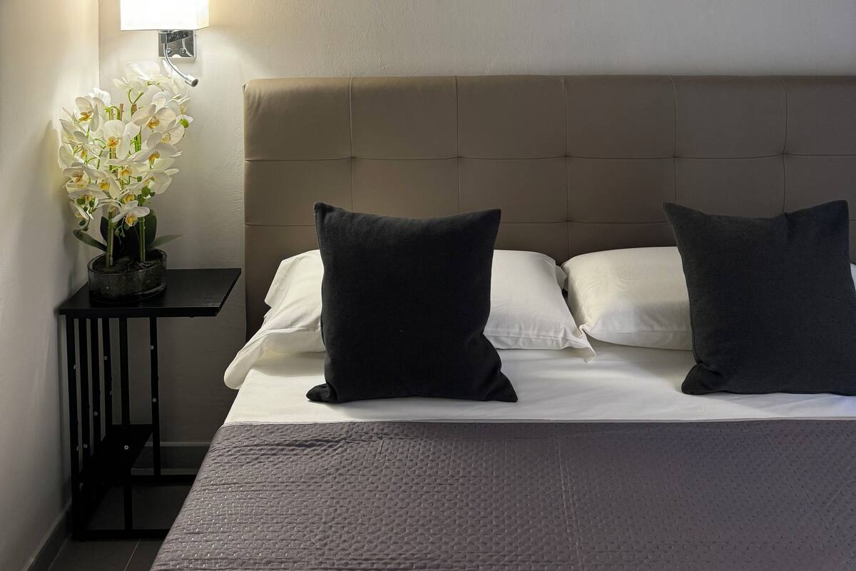 Hotelkamer met een bed, twee kussens, een nachtkastje met een lamp en een kunstwerk aan de muur.
