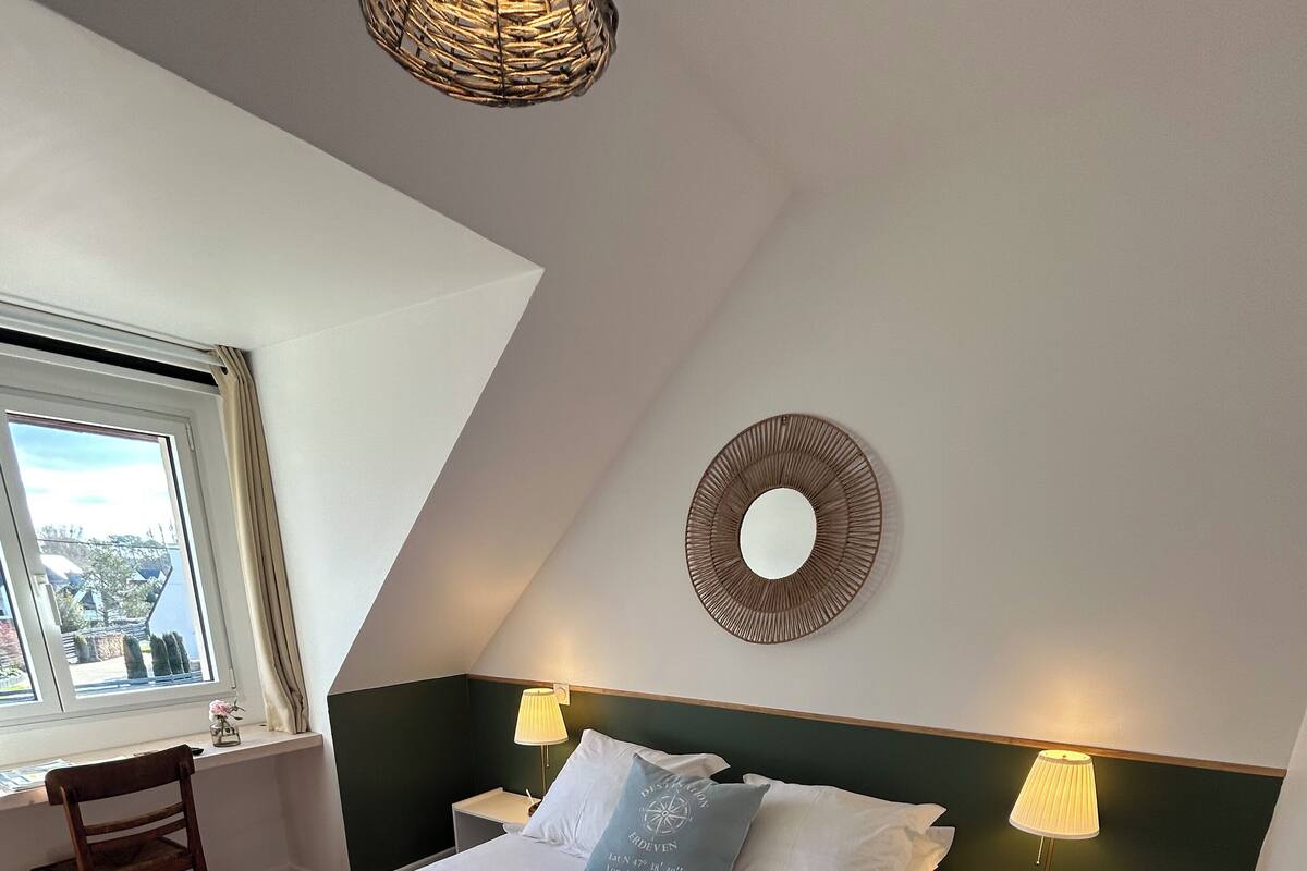 Una cama bien hecha con sábanas blancas y toallas dobladas, un espejo redondo en la pared, dos mesitas de noche con lámparas, una silla y una ventana con vistas a edificios.