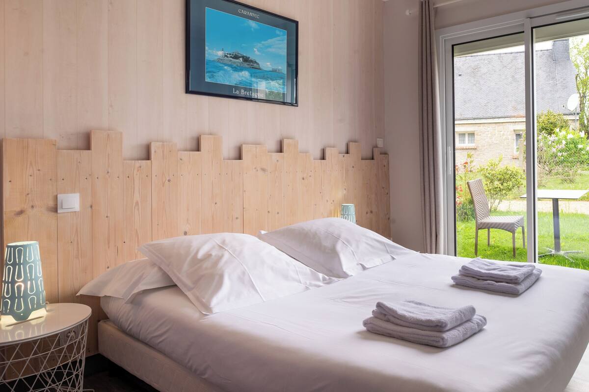 Un dormitorio con cabecera de madera, cama con ropa blanca, lámpara de noche y vistas a un jardín a través de una puerta corrediza de vidrio.