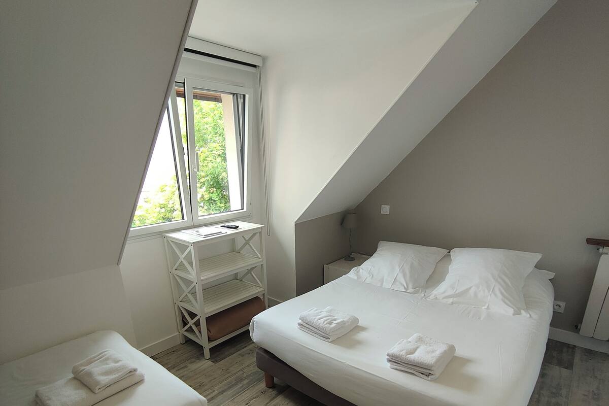 Un dormitorio con techo inclinado, una cama grande con ropa blanca, una cama más pequeña, un escritorio y una ventana con cortinas.