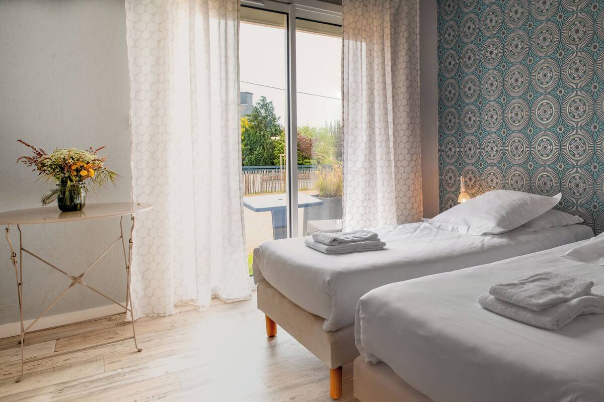 Una habitación de hotel con dos camas, una mesita con un florero y un papel tapiz estampado.