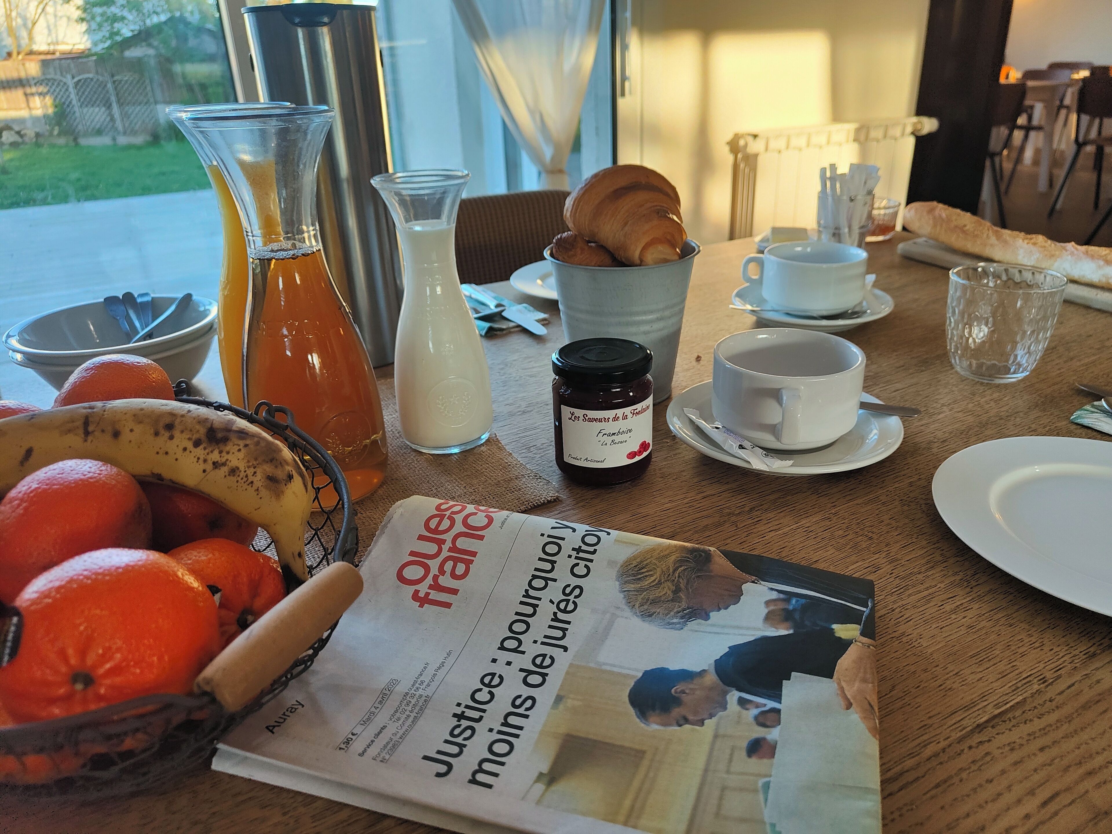 Una mesa de desayuno con cruasanes, naranjas, un periódico, una botella de jugo y un frasco de mermelada.