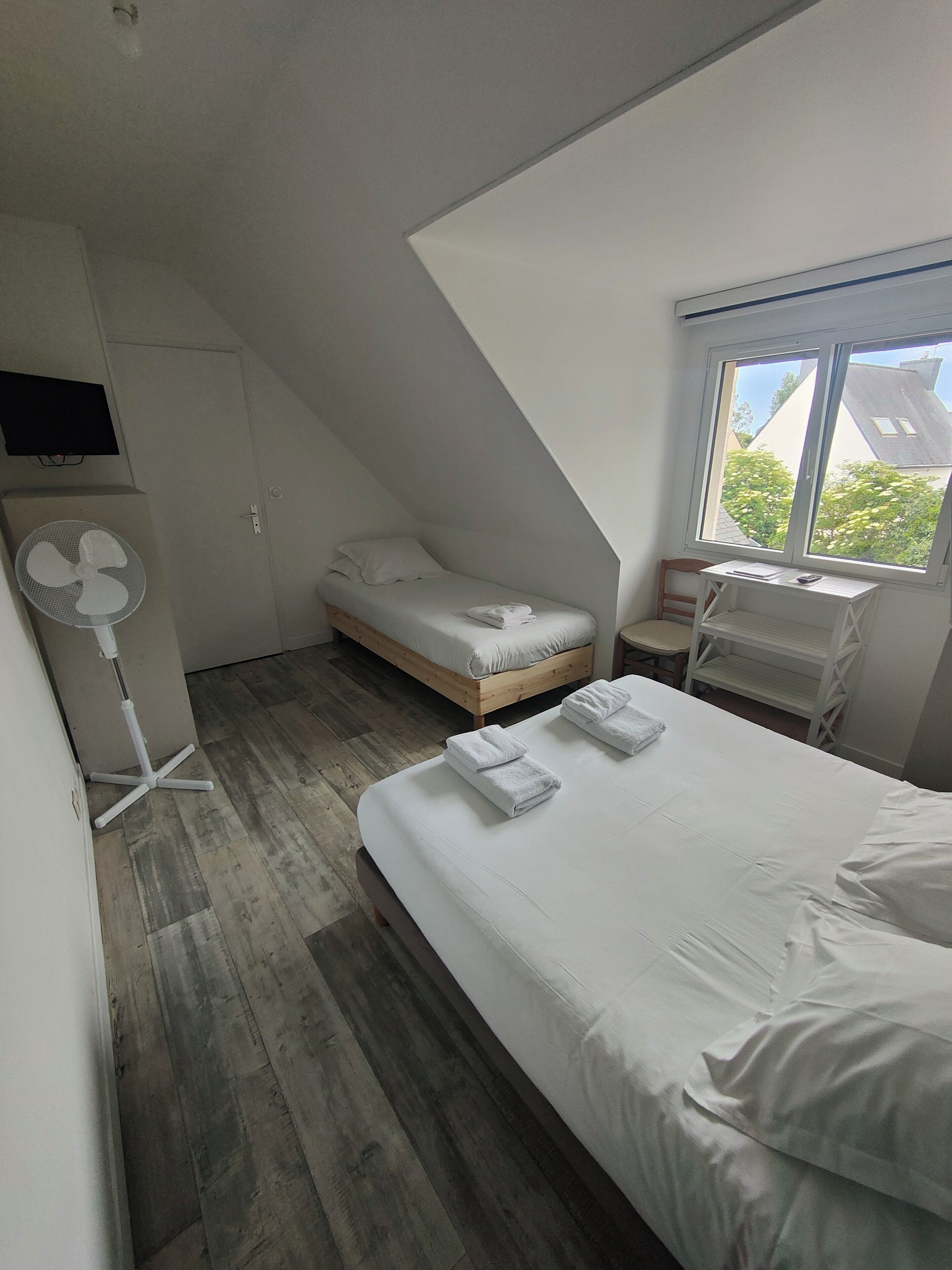 A small, modern bedroom with a sloped ceiling, a bed with white linens, a bedside table, a chair, a fan, and a window with a view of buildings and trees.