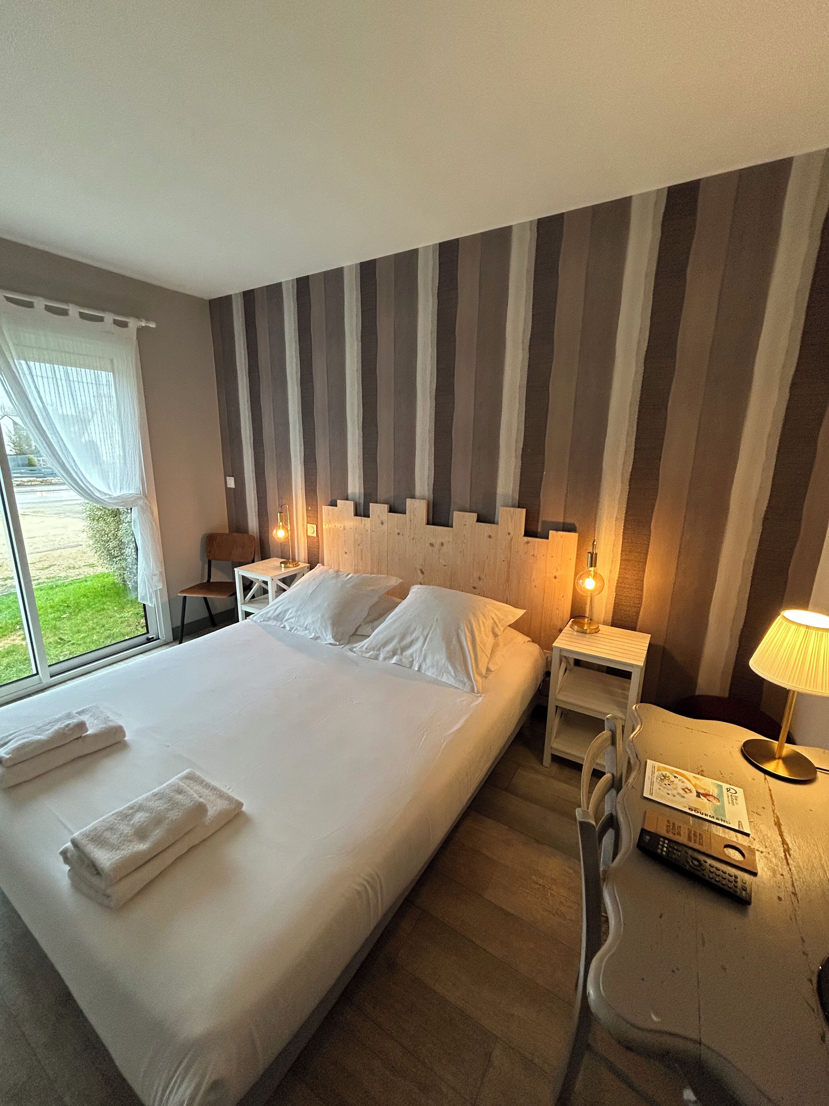 Una habitación de hotel con una cama grande, un cabecero de madera, una mesita de noche con una lámpara y vistas al exterior a través de una ventana.