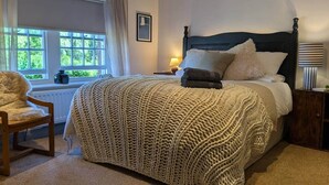 Un dormitorio con una cama grande, una silla, un ventanal con cortinas y una mesita de noche con lámpara.