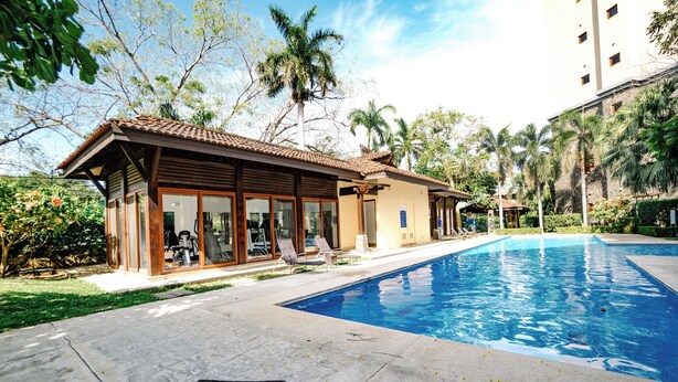 A swimming pool with a tiled deck, lounge chairs, and a building with large windows and a tiled roof.