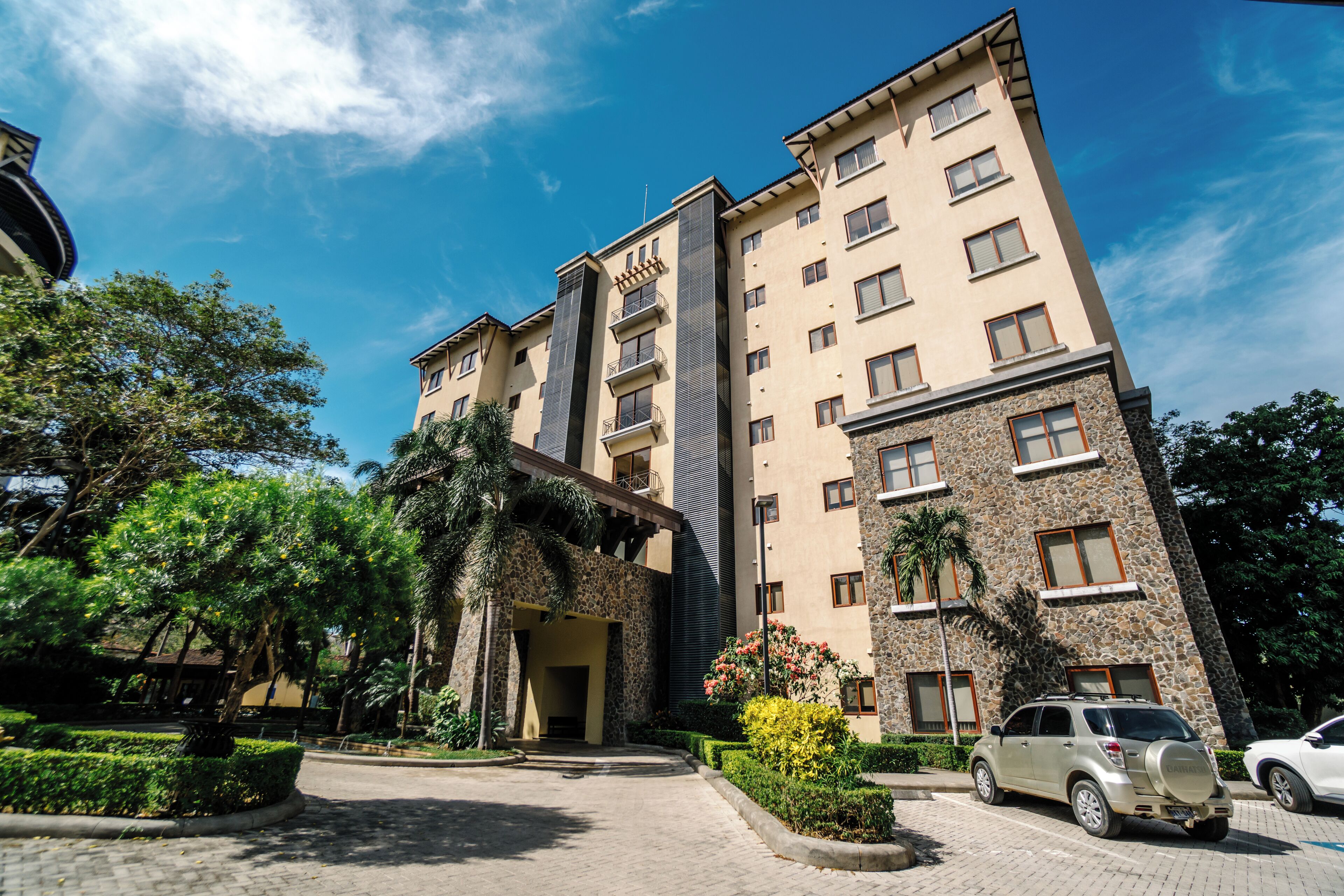 A multi-story hotel with a stone facade, a central entrance, and a parking area with cars.