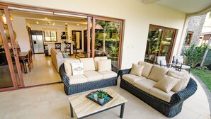 A spacious living area with two wicker sofas, a coffee table, and a sliding glass door leading to a kitchen and dining area.
