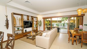 A spacious living room with a sofa, coffee table, wooden dining table, and chairs. There is a TV on a wooden stand, and a bookshelf filled with various items.
