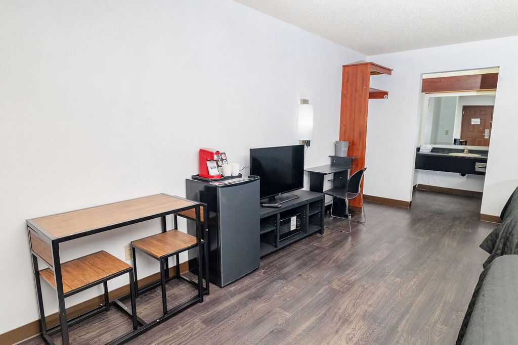 Quarto de hotel pequeno com uma escrivaninha de madeira, televisão, máquina de café e um espaço de estar aconchegante.