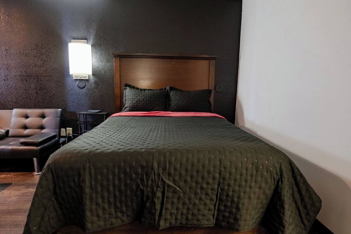 Quarto de hotel com uma cama grande, cabeceira de madeira, mesa de cabeceira, abajur e um espaço de estar com uma cadeira e uma pequena mesa.