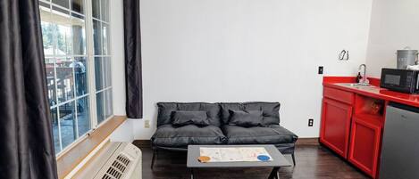 A compact living space with a black leather sofa, a small coffee table, a kitchenette with a microwave, and a window with curtains.