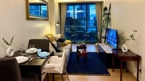 Una sala de estar moderna con sofá, mesa de comedor y televisor. Un ventanal ofrece vistas de la ciudad.