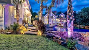 Un jardin bien entretenu, avec un chemin de pierre menant à une zone de piscine, entouré de palmiers et d’une grande maison dotée de nombreuses fenêtres.