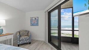Een slaapkamer met een bed, een stoel, een bureau, een schilderij en een grote schuifglazen deur met uitzicht op een strand.