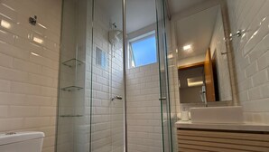 A modern bathroom with a glass-enclosed shower, white tiled walls, a wooden vanity, and a skylight. - RCM Vilas - Apartamento Sofisticado (Joinville)