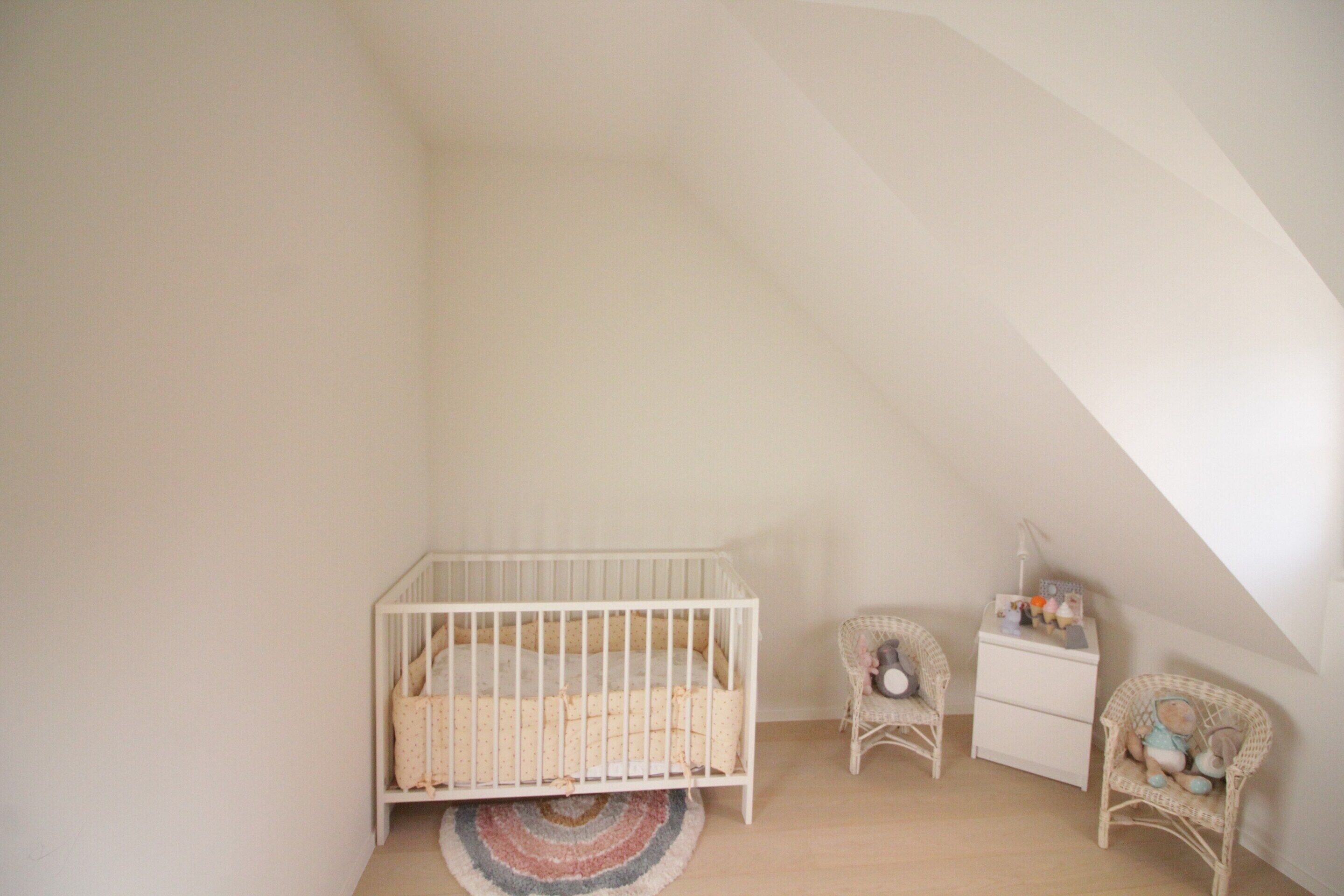 Ein Zimmer mit hohen Decken, einem weißen Babybett, einem Korbstuhl und einer kleinen Kommode.