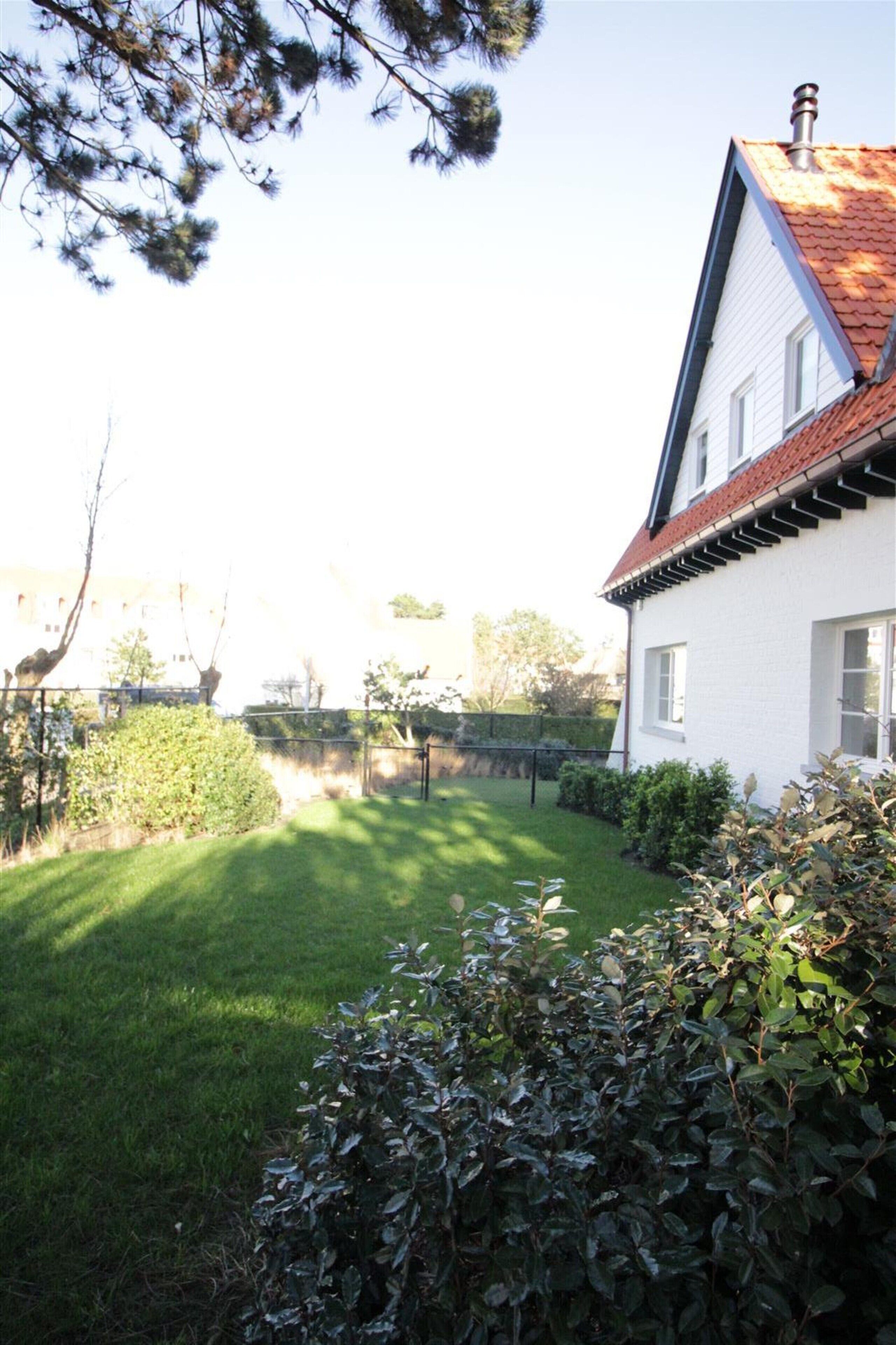 A white house with a red roof, surrounded by a well-maintained lawn and dense shrubbery.