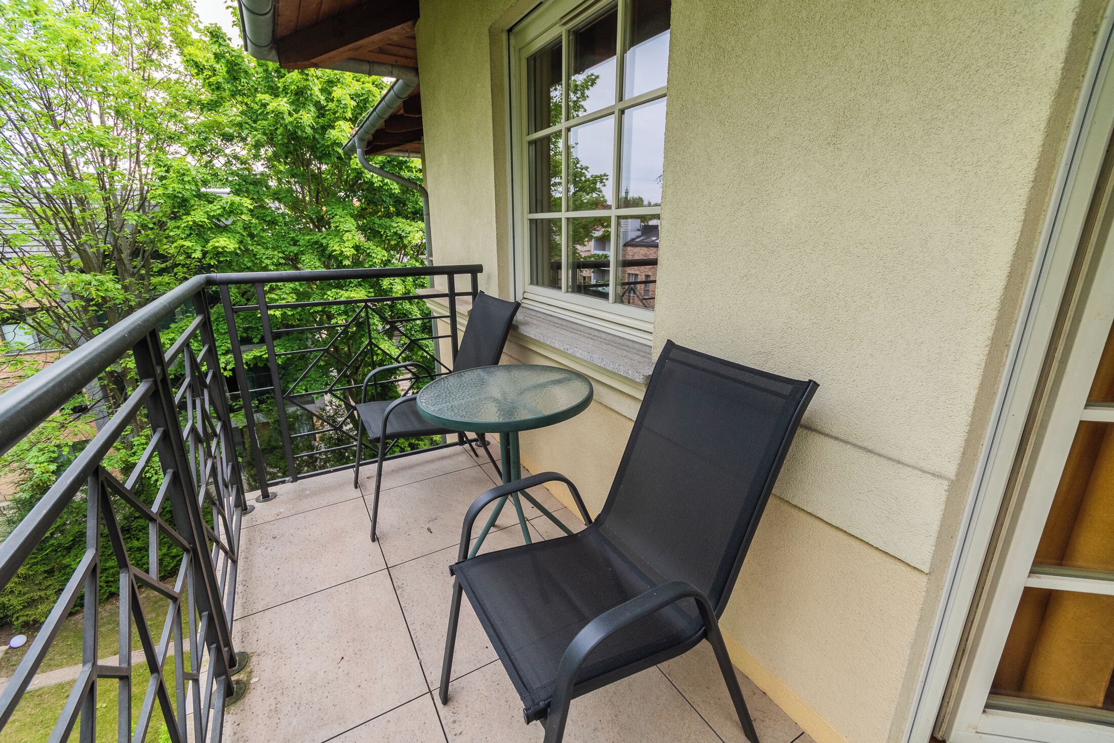 Varanda com mesa de vidro e duas cadeiras, corrimão e vista para áreas verdes e edifícios.
