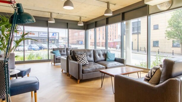 A modern lounge area with leather sofas, wooden tables, and pendant lighting. Large windows offer views of the street. - The Croft (Derby)
