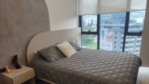 A modern bedroom with a large bed, a nightstand, and a window with a city view. - Suite XOE in front of Carolina Park - 11th floor (Quito)