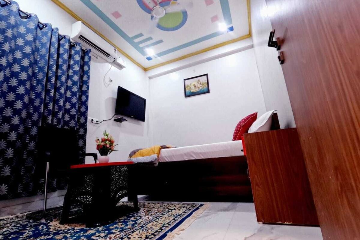 Quarto com cama, televisão, escrivaninha com cadeira, mesinha com um vaso e tapete estampado.