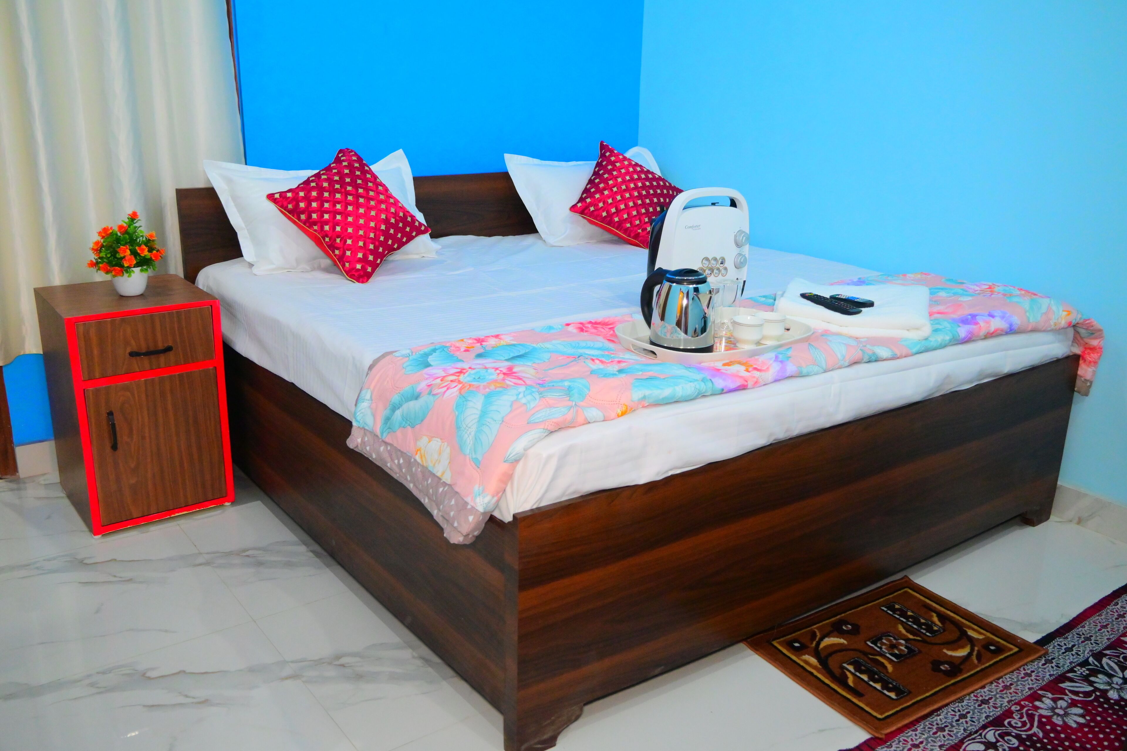 A neatly made bed with a floral bedspread, a bedside table with a red cabinet, and a coffee maker on the bed.