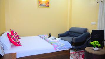 A neatly made bed with floral pillows, a bedside table with a cup and a book, a grey armchair, and a small table with a plant.