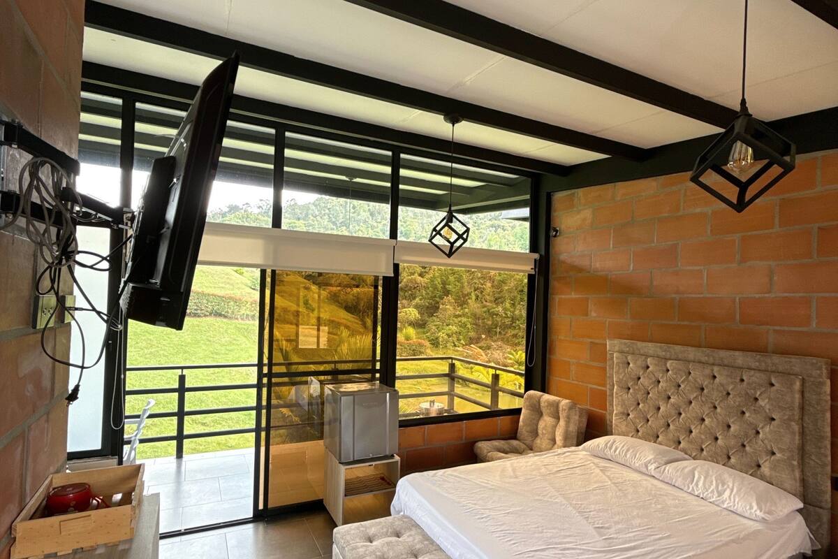 A bedroom with a large bed, a television, and a balcony with a view of greenery.