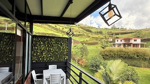 A balcony with a table and chairs, a hanging light fixture, and a view of green hills and a house. - Suites del Lago (Marinilla)