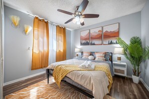 Une chambre à coucher avec un lit, un ventilateur de plafond, une table de chevet, une lampe, une plante et des œuvres d’art accrochées au mur.