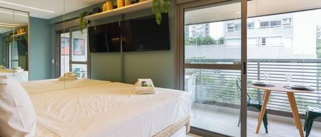 A modern hotel room with a large bed, a balcony with a table and chairs, and a view of the cityscape.