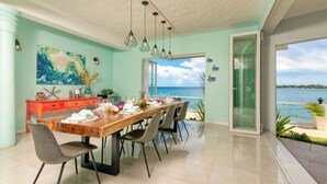 A dining area with a view of the sea, a wooden table set for six, and a teal-colored wall with a large abstract painting. - Spacious 5-bedroom villa w/ Private Beach Access 1 minute away in, St. Ann (St. Ann)