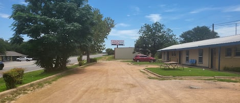 A rural road leading to a motel with a dirt driveway, a red car parked, and a large billboard in the distance.