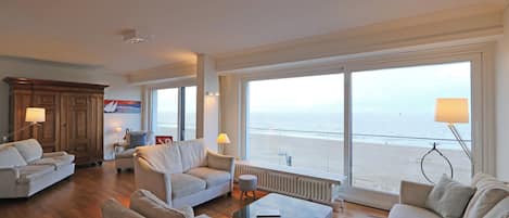 A spacious living room with a large window overlooking the beach, wooden flooring, and modern furniture.