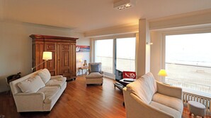 Amplia sala de estar con piso de madera, un sofá color beige, un sillón pequeño y una mesa de centro. Hay dos ventanas que ofrecen vista a la playa.