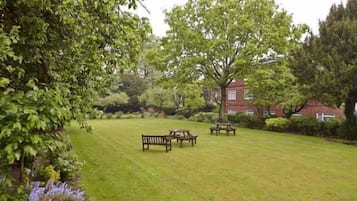 A grassy outdoor area with picnic tables and benches, surrounded by trees and a building in the background.
