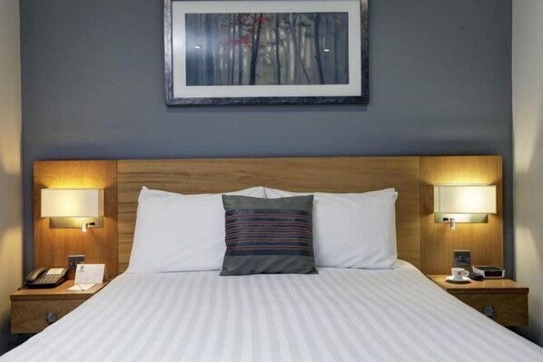 Une chambre d’hôtel avec un grand lit, deux lampes de chevet, un téléphone, une tasse et un tableau encadré au mur.