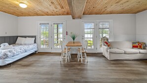 Ein geräumiges Zimmer mit Holzbalkendecke, einem Bett mit Blumenmuster-Überwurf, einem Esstisch mit Stühlen, einer Couch und großen Fenstern, die viel natürliches Licht hereinlassen.