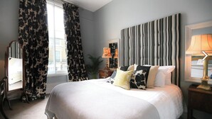 A bedroom with a large bed, a mirror, a bedside table with a lamp, and a window with curtains. - Crown and Sceptre (London)